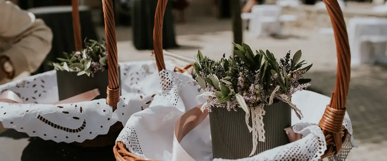Zwei geflochtene Körbe mit Lavendel und Olivenzweigen, bereit für eine kroatische Hochzeit. Weiße Spitzendeckchen und rosa Bänder zieren die Körbe.