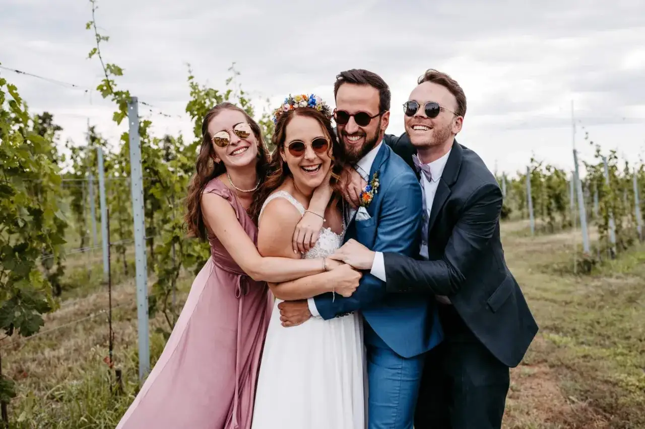 Gl&uuml;ckliches Brautpaar mit seinen Hochzeit Trauzeugen in einem Weinberg, alle tragen Sonnenbrillen und umarmen sich fr&ouml;hlich.