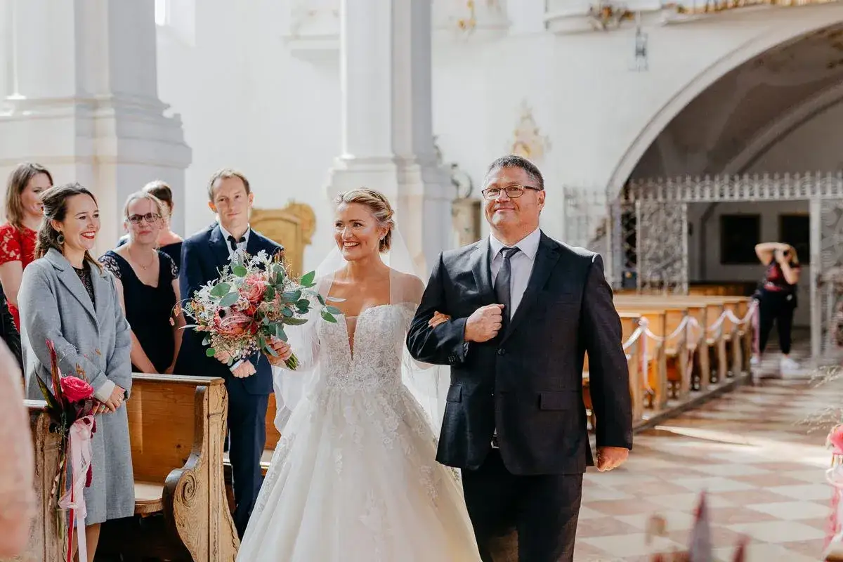 Ein Vater und seine Tochter gehen Arm in Arm durch die Kirche. Sie l&auml;cheln, ein Moment des Stolzes und der Freude, der an einen vater tochter tanz erinnert.