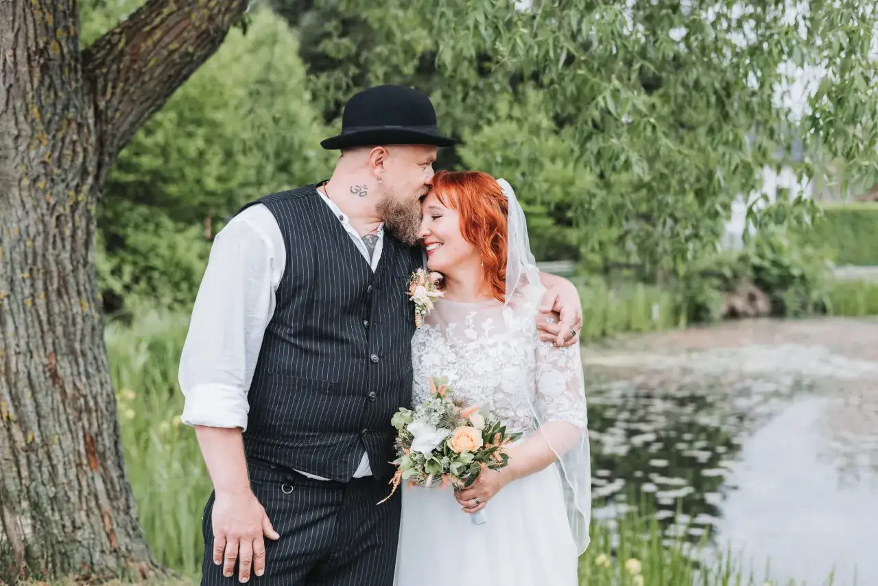Ein Brautpaar umarmt sich liebevoll vor einem Teich. Der Br&auml;utigam tr&auml;gt einen Hut und eine Weste, die Braut ein Spitzenkleid. Ein sch&ouml;ner Spruch f&uuml;r die Ehe.