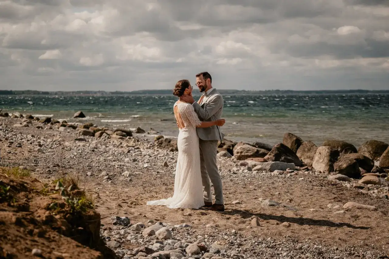 Ein Brautpaar genie&szlig;t seine Hochzeit am Strand zu zweit. Die Braut tr&auml;gt ein Spitzenkleid, der Br&auml;utigam einen grauen Anzug. Das Meer und der Himmel sind im Hintergrund zu sehen.