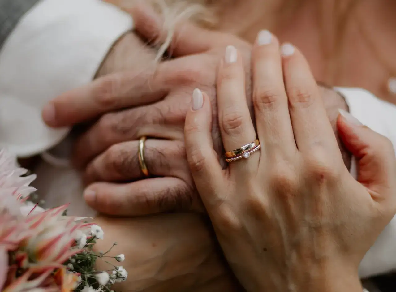 Zwei H&auml;nde, die sich halten. Ein Mann tr&auml;gt einen goldenen ehering, eine Frau tr&auml;gt zwei zarte Ringe, einer davon mit einem Stein.