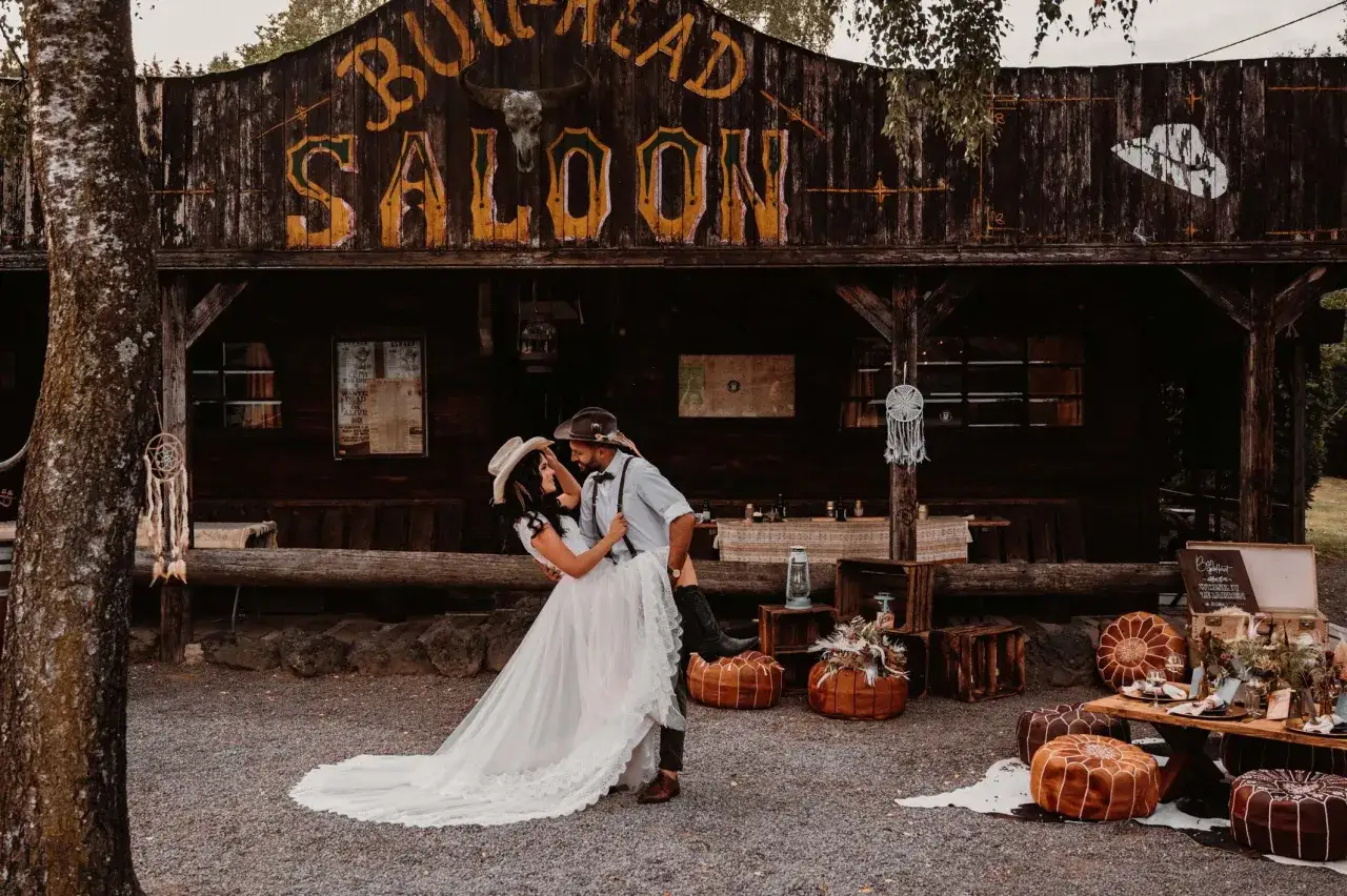 Ein Brautpaar feiert seine western hochzeit vor einem Saloon. Sie tanzen ausgelassen, umgeben von rustikaler Deko.
