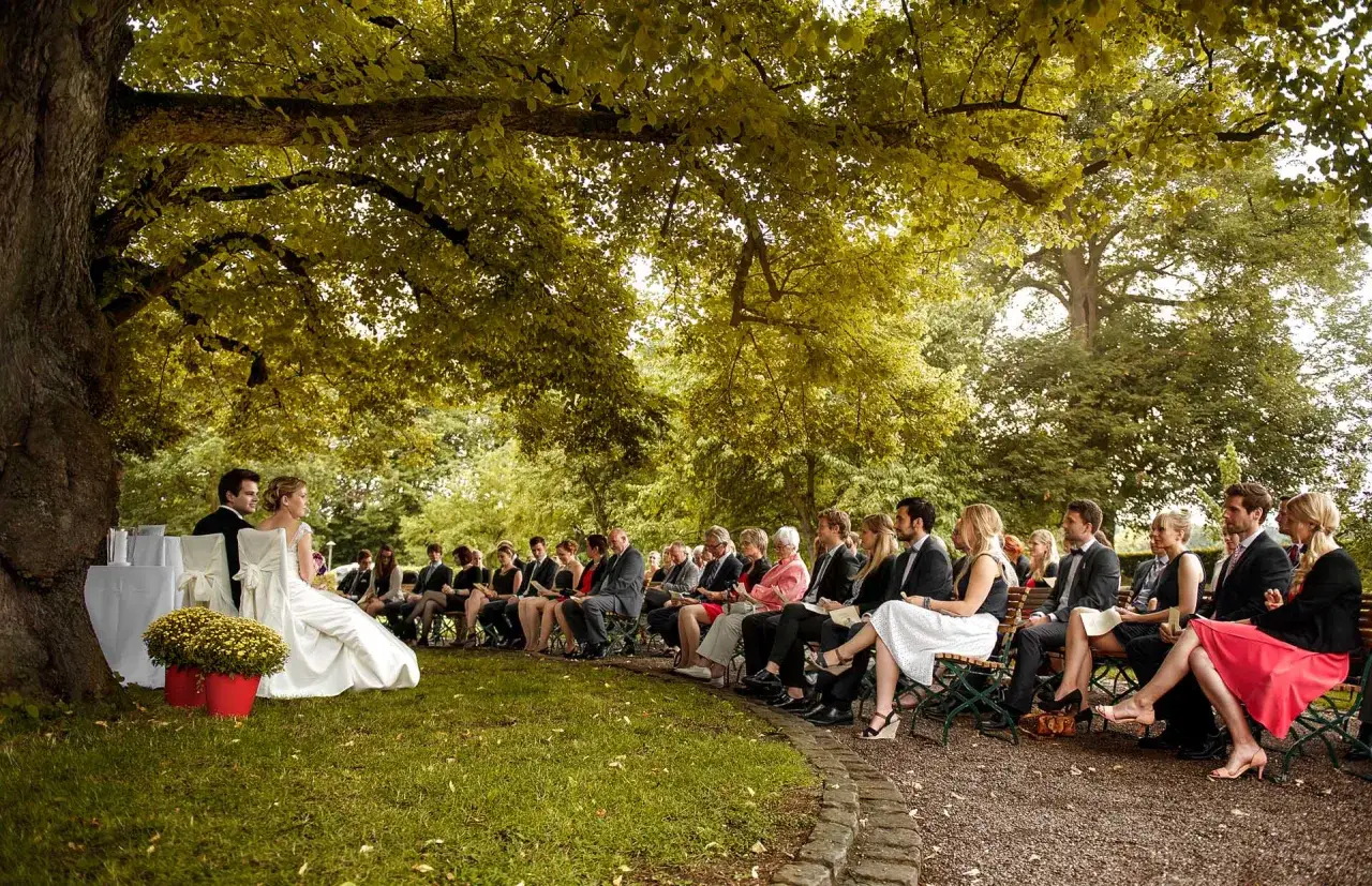 Hochzeitspaar bei freier Trauung mit Standesbeamten unter einem Baum. G&auml;ste sitzen auf B&auml;nken im Kreis.