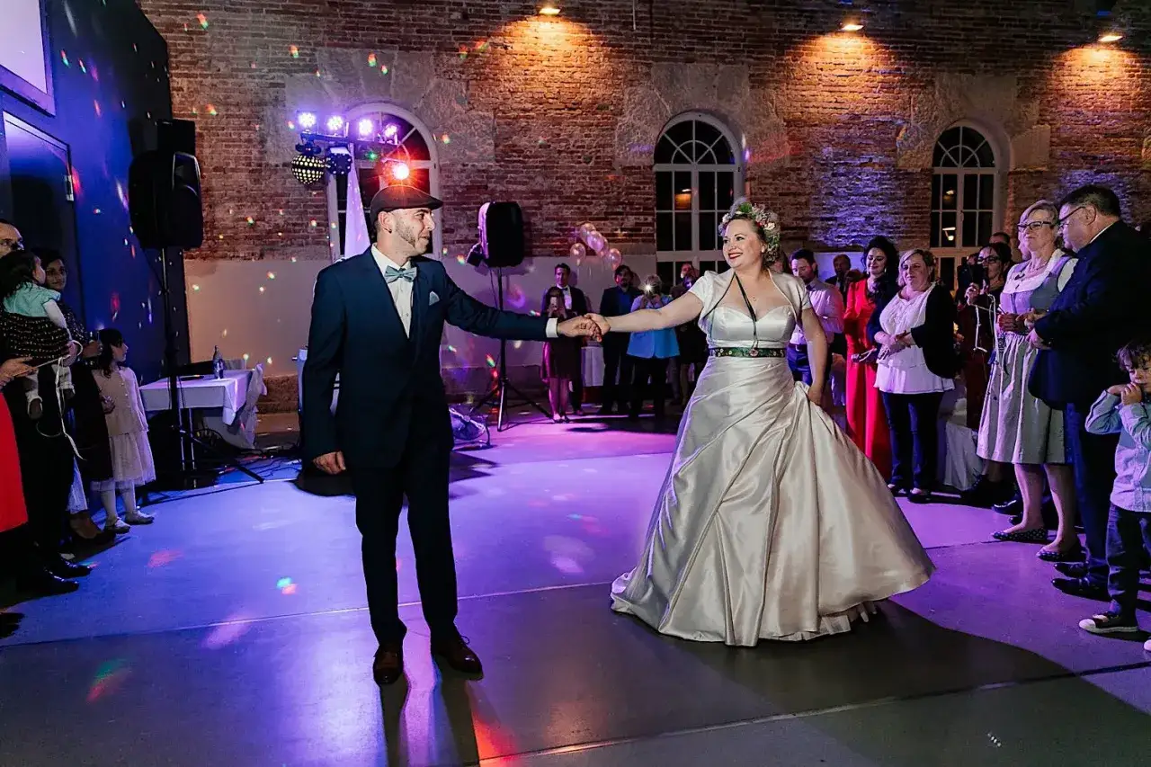 Brautpaar &uuml;bt den Hochzeitstanz. Sie halten H&auml;ndchen und l&auml;cheln sich an, w&auml;hrend G&auml;ste zusehen. Bunte Lichter tanzen auf dem Boden.