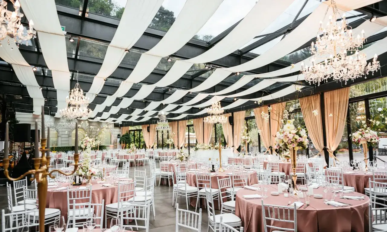 Festlich gedeckte Tische mit rosa Tischdecken und wei&szlig;en St&uuml;hlen unter Kronleuchtern. Eine elegante Location, die sich perfekt f&uuml;r eine Hochzeit eignet, bei der man auch an eine Hochzeit Versicherung denken sollte.