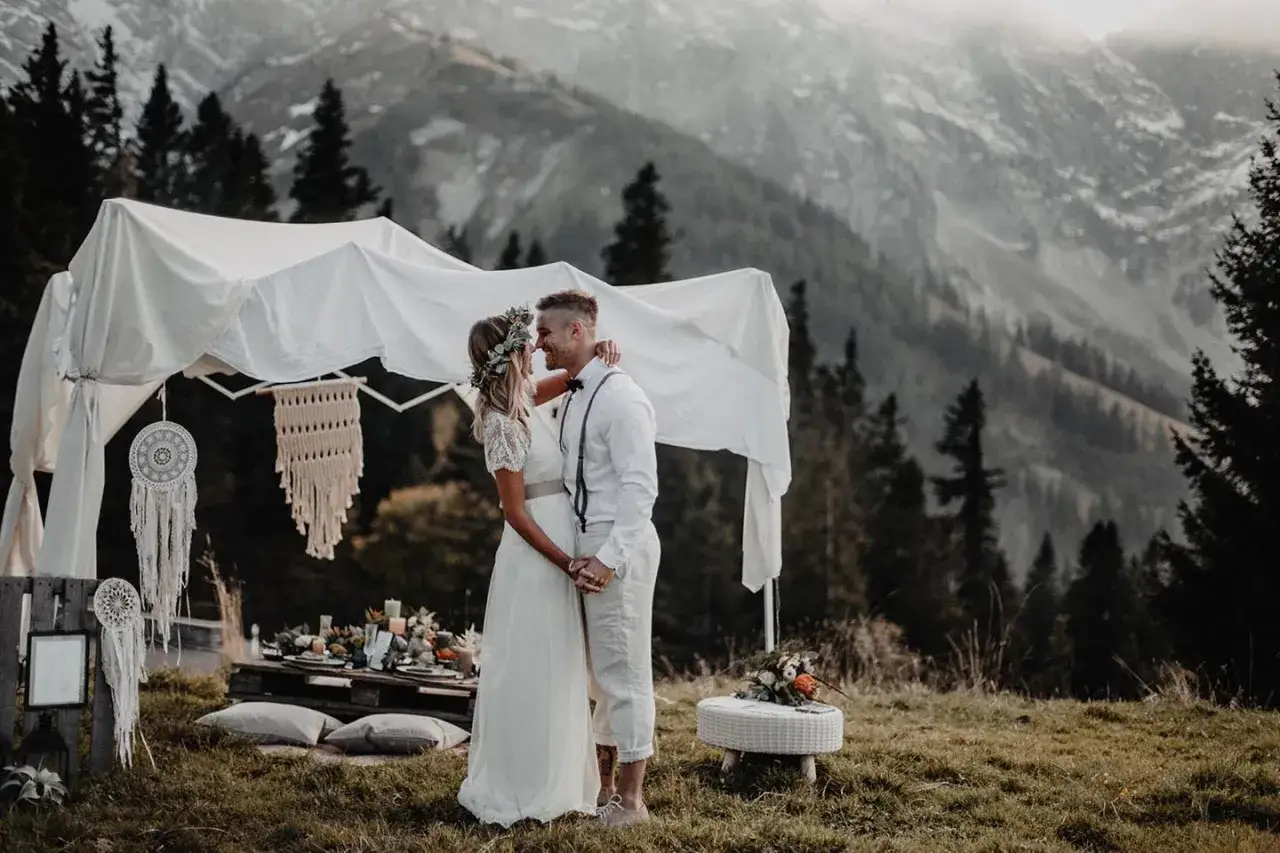 Ein Brautpaar umarmt sich unter einem Baldachin, umgeben von der majest&auml;tischen Kulisse einer Hochzeit in den Bergen.