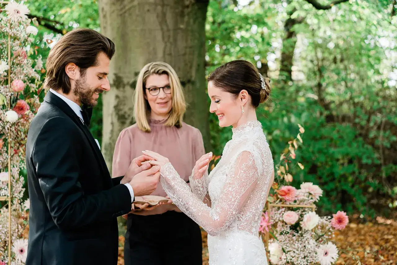 Ein Mann im Anzug steckt seiner Braut, die ein Spitzenkleid tr&auml;gt, den Ring an. Eine Frau im rosa Oberteil beobachtet die emotionale Rede beim Standesamt.