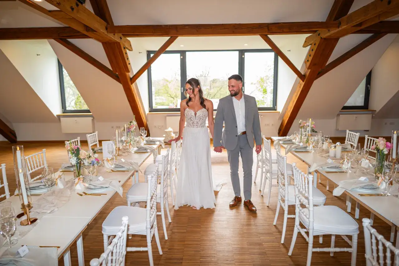 Brautpaar schreitet durch den Raum einer minimalistischen Hochzeit. Wei&szlig;e St&uuml;hle und gedeckte Tische mit Kerzen und Blumen schaffen eine elegante Atmosph&auml;re.