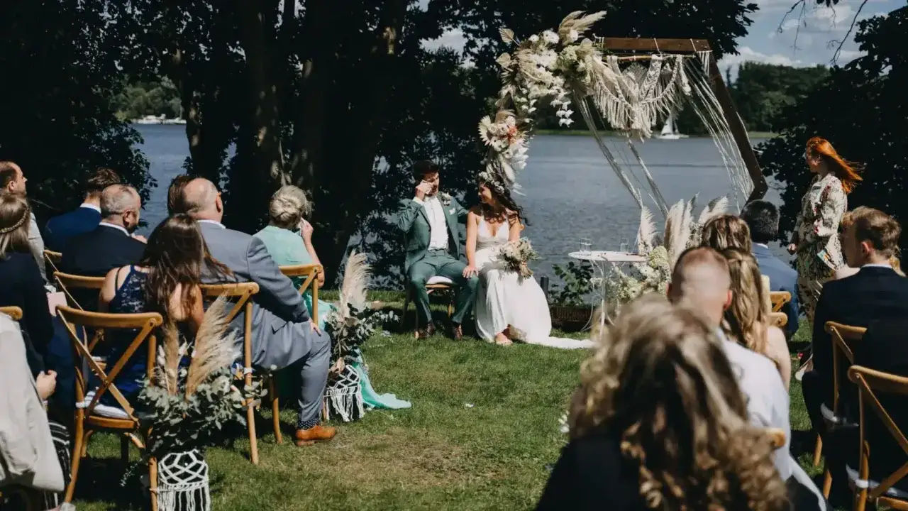 Hochzeit am See mit viel greenery. Brautpaar sitzt unter einem Bogen, G&auml;ste auf St&uuml;hlen.