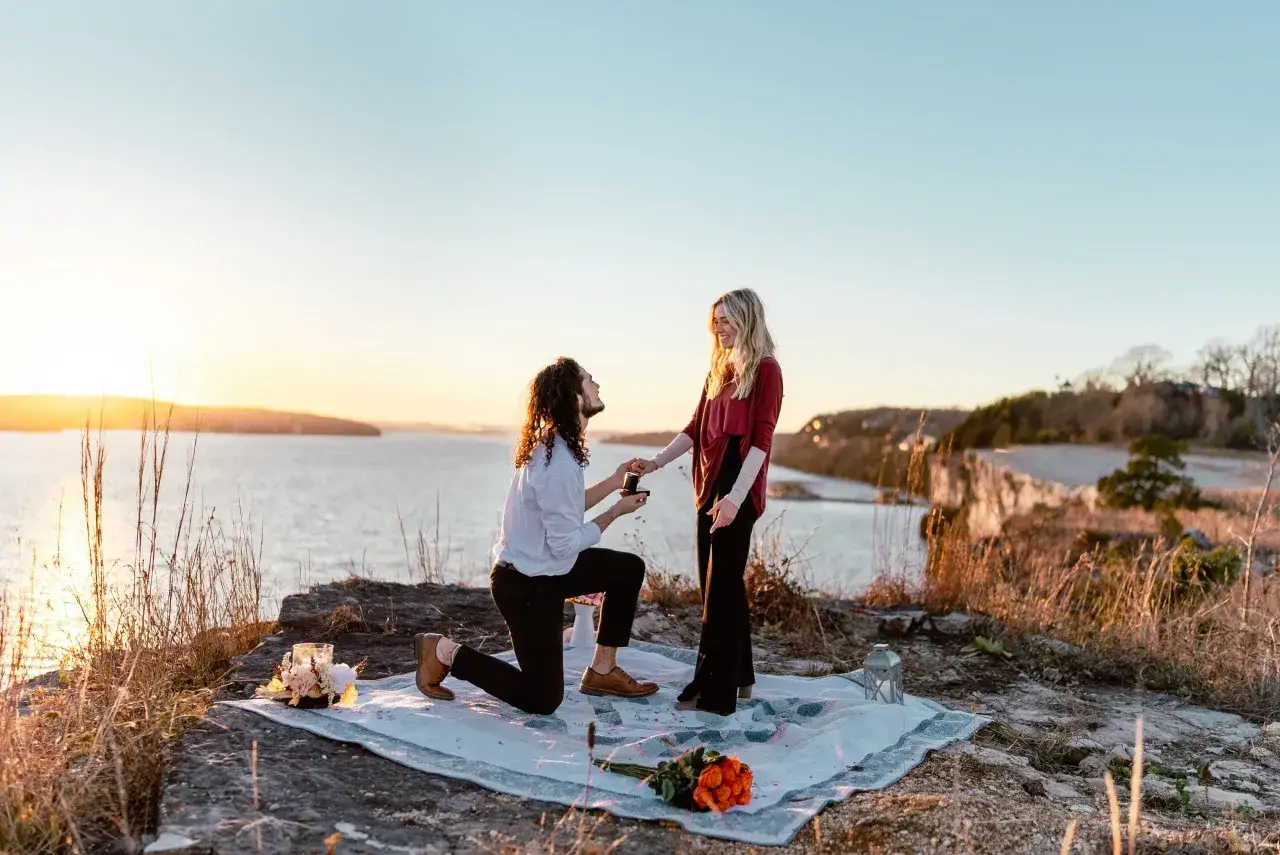Mann kniet mit Ring vor Frau nieder. Romantische Heiratsantrag Ideen am See bei Sonnenuntergang.