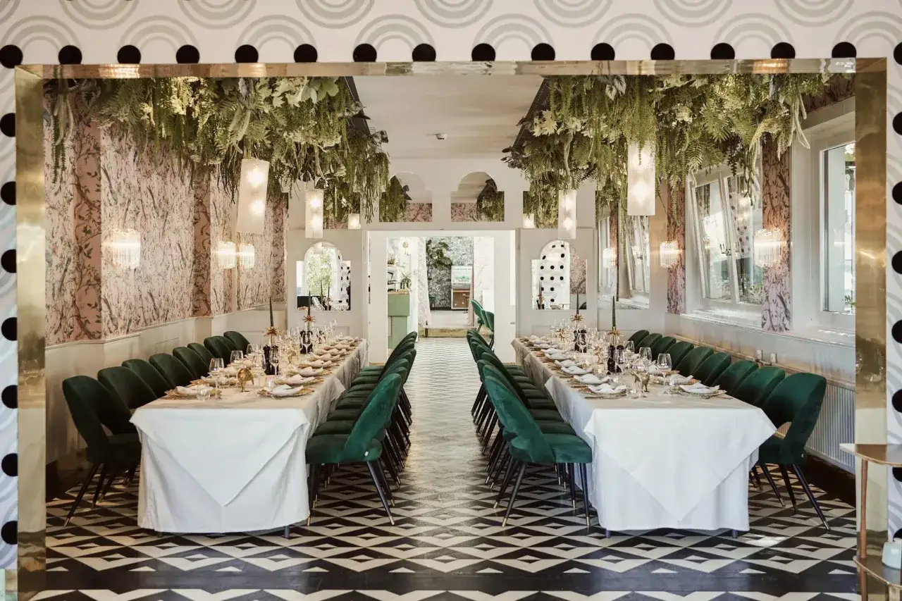 Ein Festsaal mit zwei langen Tafeln, geschmückt mit üppigem Grün von der Decke. Grüne Samtstühle säumen die Tische, bereit für eine Hochzeit mal anders feiern.