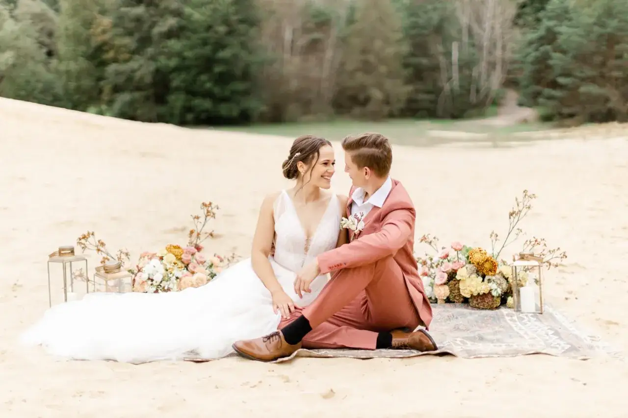 Brautpaar sitzt auf einer Decke im Sand, umgeben von Blumen. Der Bräutigam trägt einen korallenfarbenen Anzug. Ein wunderschöner Anblick, der den besten Monat zum Heiraten widerspiegelt.