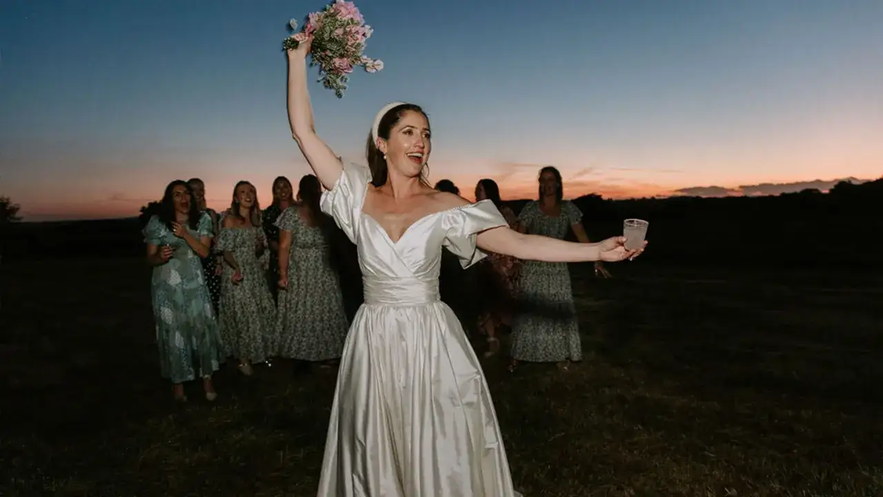 Braut wirft Blumenstrauß bei einer englischen Hochzeit im Abendlicht. Brautjungfern warten gespannt.