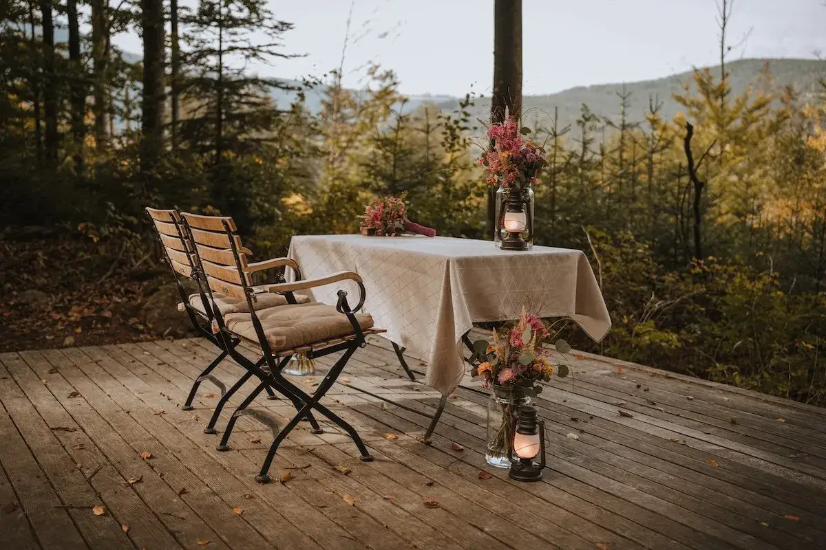 Zwei St&uuml;hle und ein Tisch mit Blumen und Laternen auf einer Holzterrasse im Wald. Ein romantischer Ort, um heimlich heiraten zu wollen.