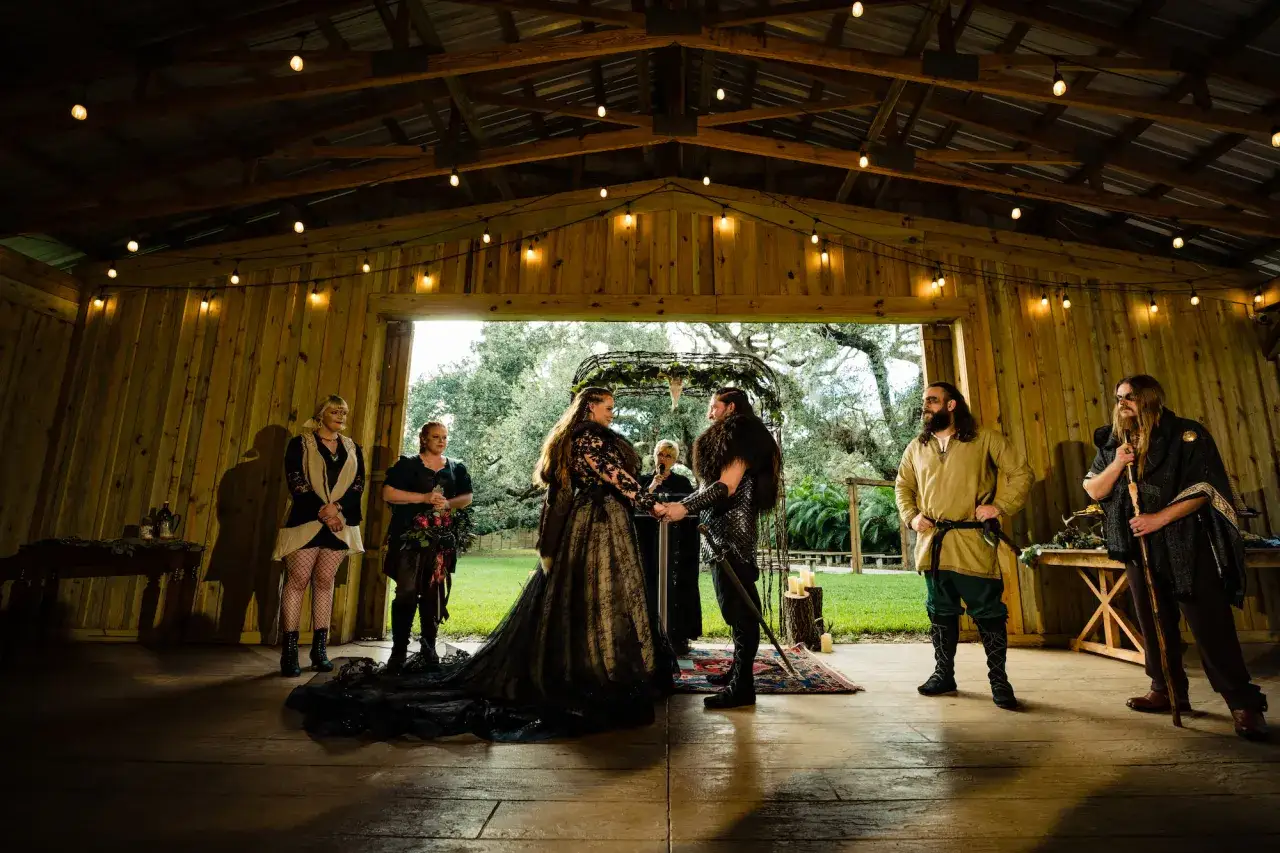Brautpaar bei einer Wikinger Hochzeit im rustikalen Holzpavillon. Sie halten H&auml;ndchen vor dem Altar, umgeben von G&auml;sten in historischen Kost&uuml;men.