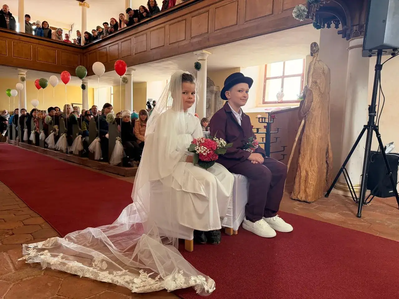 Kinder in Hochzeitskleidung sitzen auf einer Bank in der Kirche. Ein Junge tr&auml;gt einen Anzug und Hut, das M&auml;dchen ein Brautkleid. Eine klassische Hochzeit mit G&auml;sten auf den B&auml;nken.