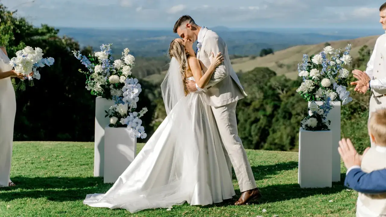 Ein Brautpaar k&uuml;sst sich bei einer modernen Hochzeit im Freien. Der Br&auml;utigam tr&auml;gt einen hellen Anzug, die Braut ein wei&szlig;es Kleid. Blumenarrangements s&auml;umen den Weg.