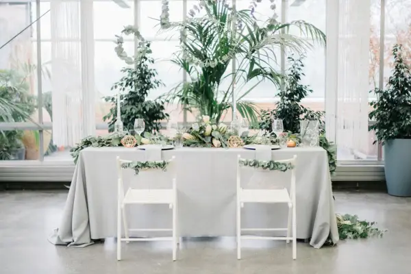 Ein festlich gedeckter Tisch für eine greenery hochzeit, umgeben von üppigem Grün. Zwei weiße Stühle mit Eukalyptus-Girlanden stehen bereit.