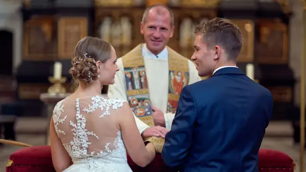 Ein glückliches Paar bei einer christlichen Hochzeit, umgeben von der feierlichen Atmosphäre einer Kirche. Der Bräutigam im blauen Anzug und die Braut im Spitzenkleid blicken sich an, während der Pfarrer sie segnet.