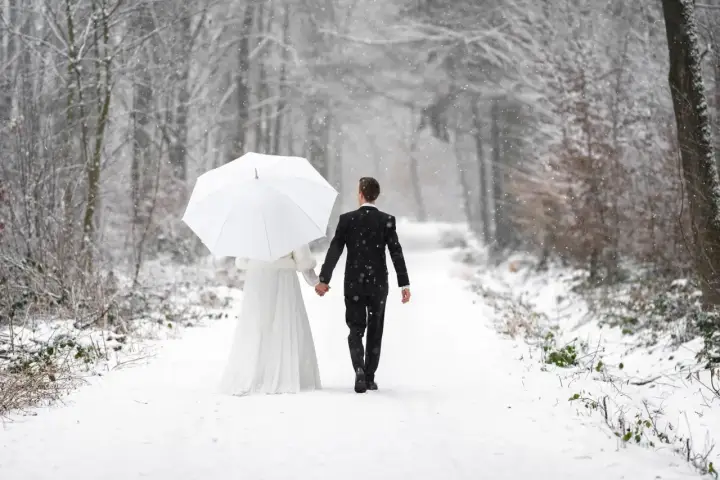 Ein Brautpaar geht Hand in Hand unter einem weißen Schirm durch einen verschneiten Wald. Vielleicht ist dies der beste Monat zum Heiraten für alle, die eine romantische Winterhochzeit wünschen.